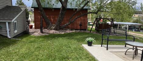 A backyard with a wooden cabin, a swing set, and a patio area with a table and chairs.