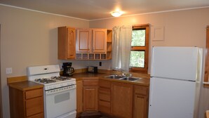 A compact kitchen with wooden cabinets, a white stove, a refrigerator, and a window with curtains.