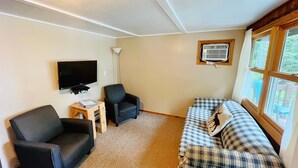 A cozy living room with a flat-screen TV, two black armchairs, a small wooden side table, and a plaid sofa.