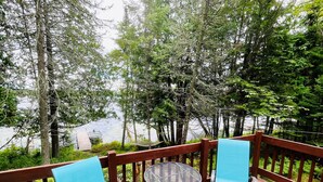 A wooden deck with two blue chairs and a small round table, overlooking a lake with trees.