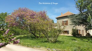 A rustic stone building with a tiled roof, surrounded by lush greenery and blooming trees.