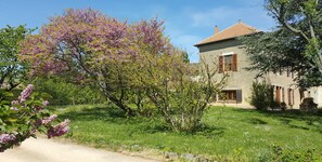 A rustic stone building with a tiled roof, surrounded by lush greenery and blooming trees.