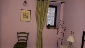 Een kamer met een stoel, een tafeltje met een lamp, een raam met behang gordijnen en een ingelijste foto aan de muur.