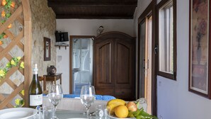 A dining area with a table set for four, a stone wall, wooden ceiling beams, a cabinet, and a window. - Apartment in studio villa called Vulcano sleeps 2 (REITANO)