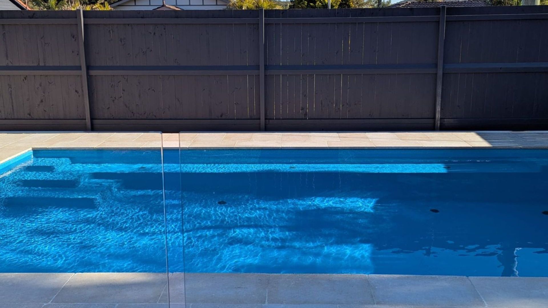 Ein rechteckiger Pool mit klarem, blauen Wasser, umgeben von einer gefliesten Terrasse und einem Holzzaun.