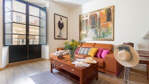 Un salon chaleureux avec une table basse en bois, un canapé agrémenté de coussins, une grande fenêtre donnant sur des immeubles et un tableau accroché au mur.