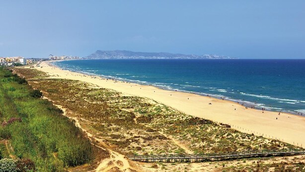 A coastal beach with a sandy shore, a wooden walkway, and buildings in the background. - Camping la Naranja (Gandia)