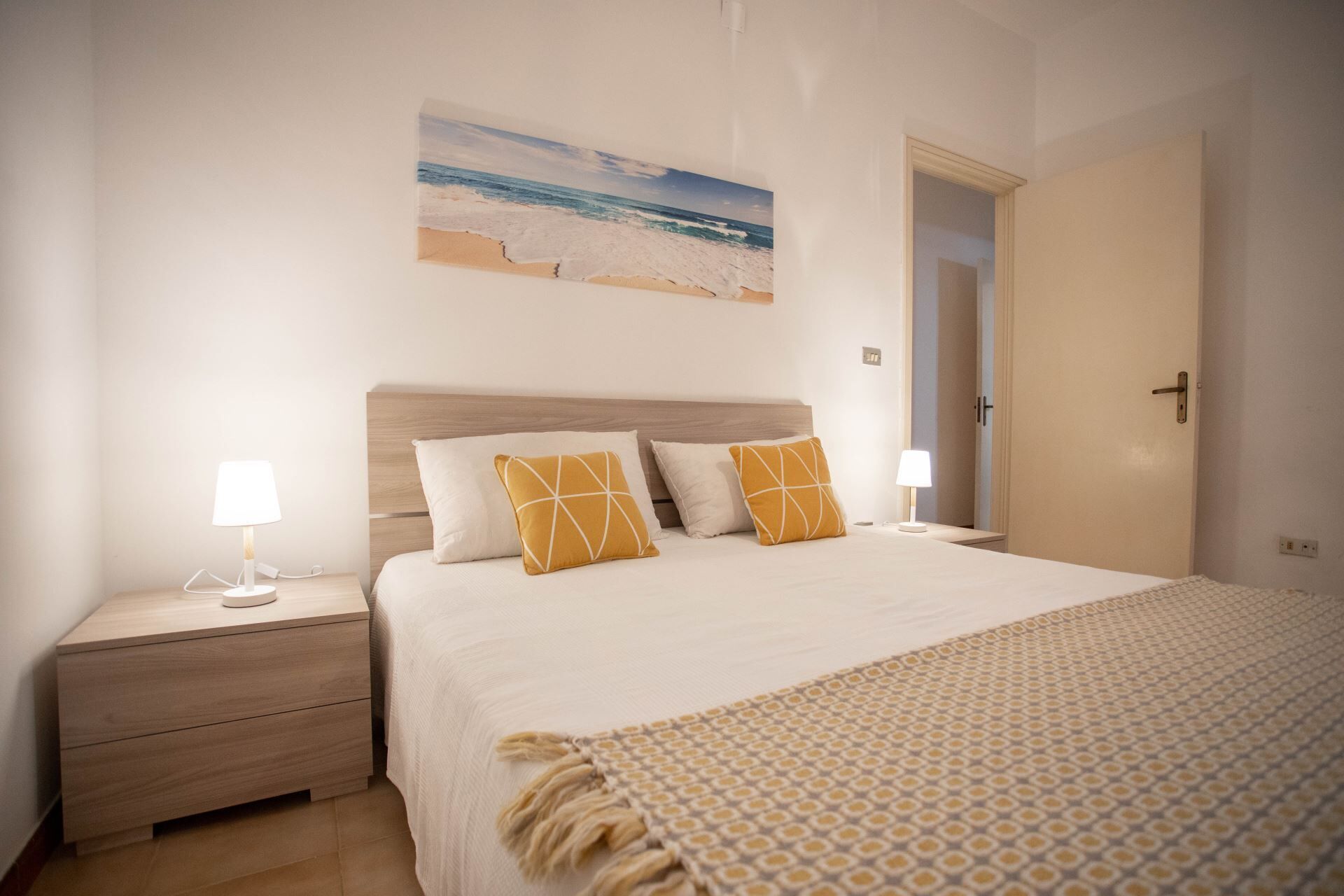 Une chambre à coucher comprenant un lit, deux tables de chevet, une commode, une porte et une œuvre d’art fixée au mur représentant une scène de plage.