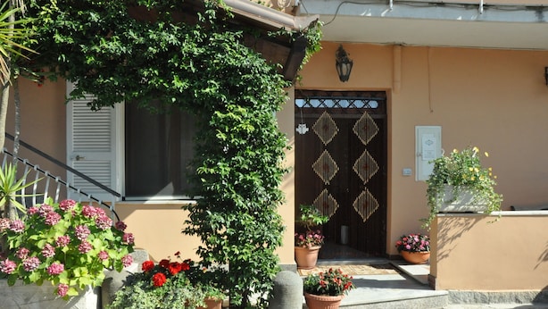 A building with a brown facade, a black door with diamond patterns, and potted plants with flowers. - Victoria (Ercolano)