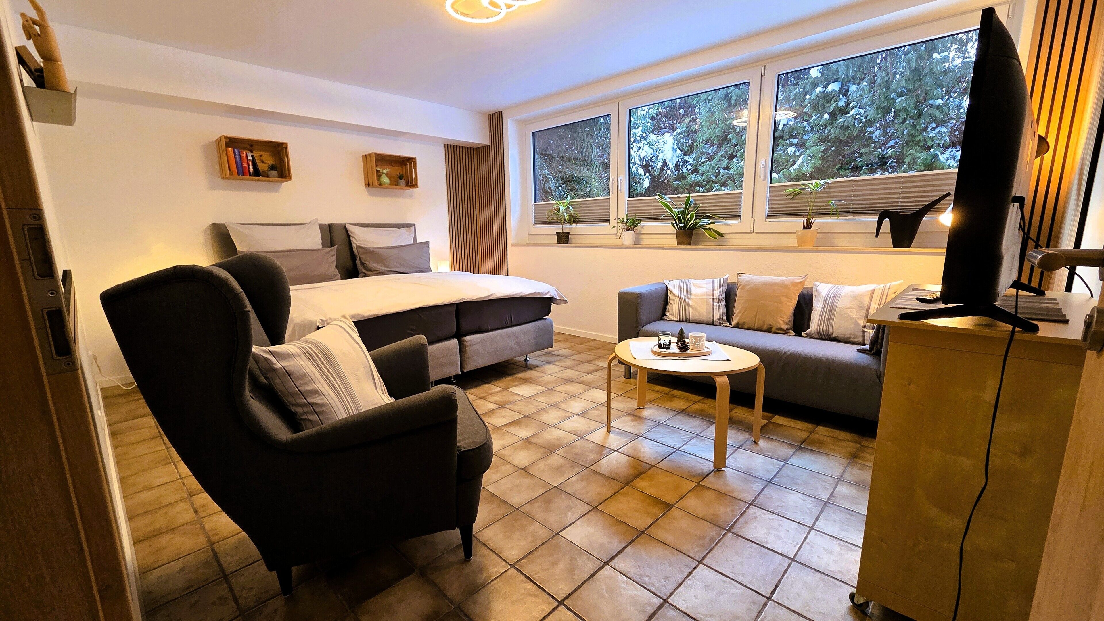 Un salon moderne avec un canapé, un fauteuil et une table basse. On y trouve un lit avec des oreillers et un téléviseur posé sur un meuble.