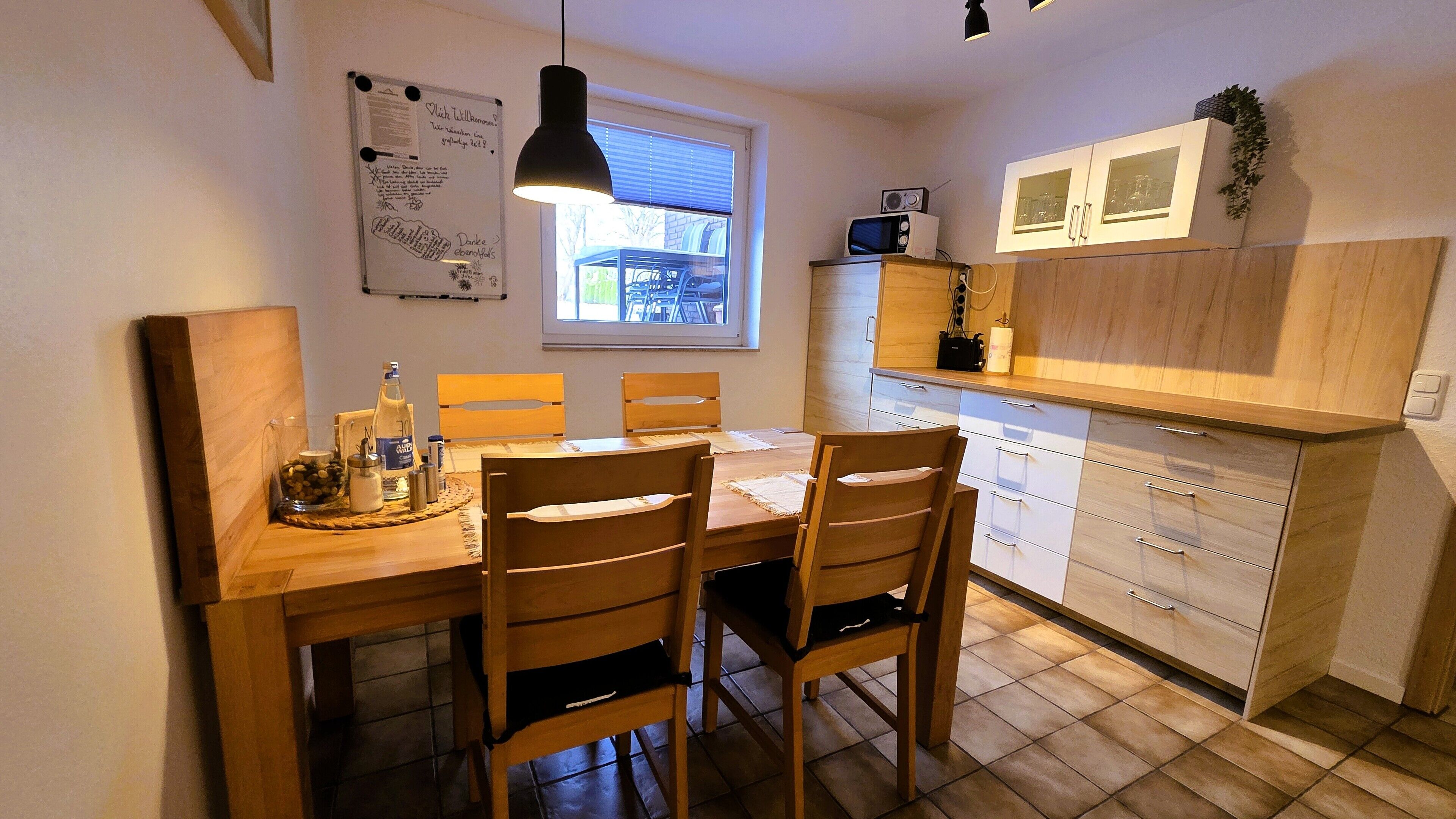 A compact kitchen with a dining table, four wooden chairs, a hanging pendant light, and a window with blinds.