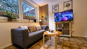 A living room with a grey sofa, a small wooden table, a TV on a wooden stand, and a window with blinds. - 3 stars: Apartment on the ground floor, also suitable for wheelchair users (Hamburg)