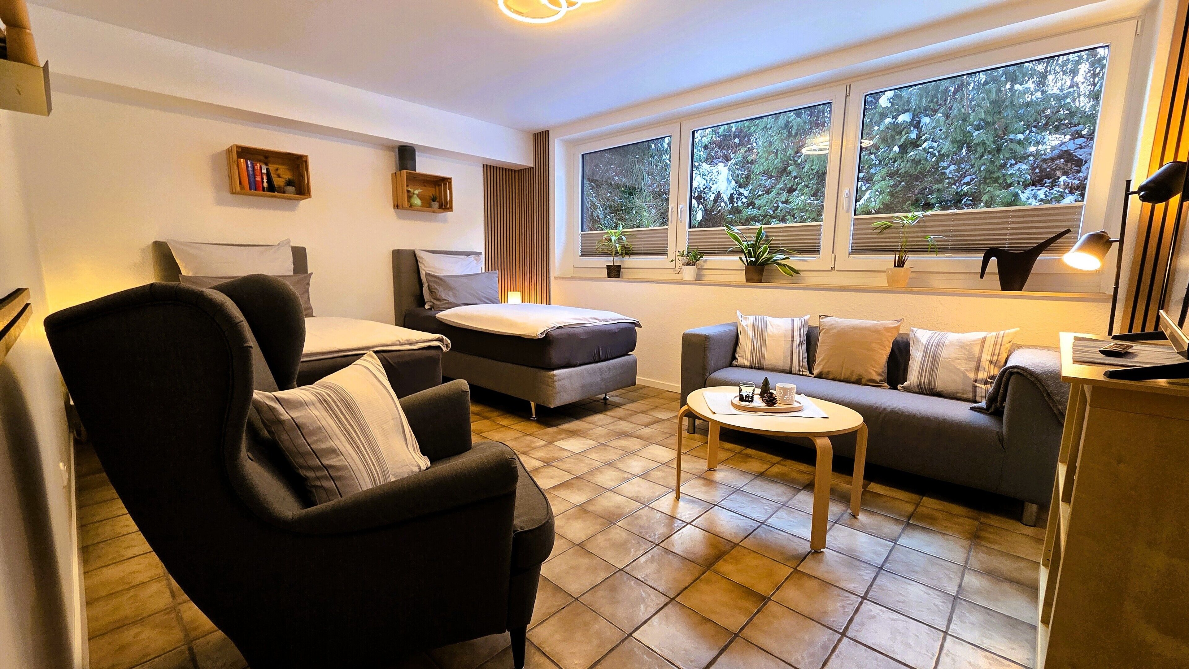A modern living room with a sofa, armchair, and coffee table. There are two windows with blinds, a lamp, and a small shelf with books.