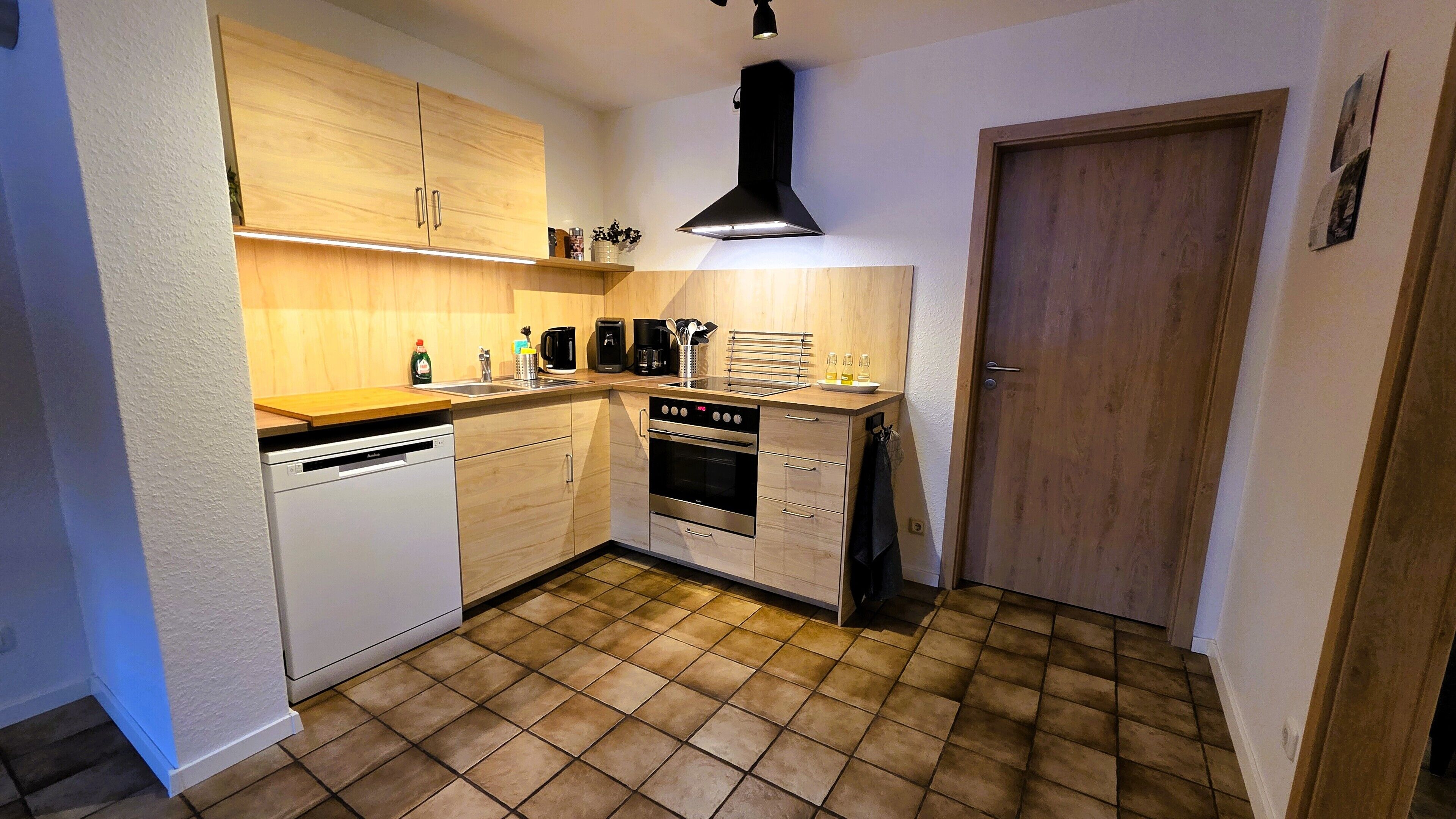 Eine kompakte Küche mit Holzschränken, einem Geschirrspüler, einem Backofen und einer schwarzen Dunstabzugshaube.