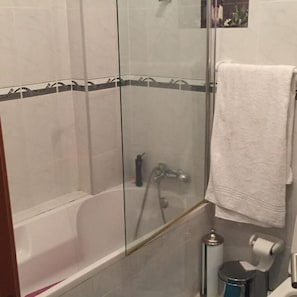 Zona de ducha con paredes de vidrio, una bañera, una toalla colgada en la pared y un cesto de basura.