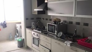 Una cocina con microondas, estufa y armarios. Hay una lavadora en la esquina y un cubo de basura.