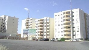 Un edificio residencial de varios pisos con balcones, cielo despejado y coches aparcados en primer plano.