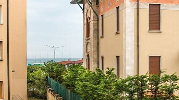 A narrow street with buildings on either side, featuring a mix of architectural styles and a view of the sea in the distance.