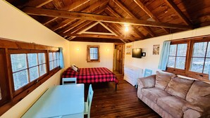 A cozy room with a wooden ceiling, a bed with a red and black checkered bedspread, a white dresser, a brown sofa, and a table with chairs.