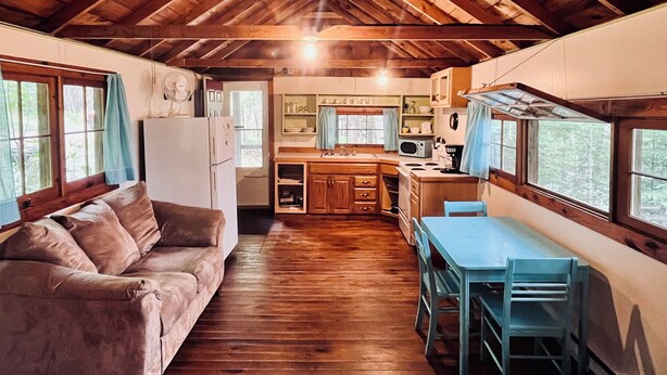A cozy cabin interior with a wooden ceiling, a kitchenette, a dining area, and a living room with a sofa.