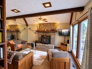 A cozy living room with a stone fireplace, a ceiling fan, and a large window with curtains. - Timberline (Ruidoso)