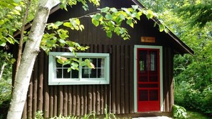 Une petite cabane en bois avec une porte rouge et une fenêtre, entourée d’arbres.