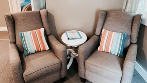 Deux fauteuils ornés de coussins à rayures, une petite table ronde avec un livre, et une fenêtre avec des rideaux.