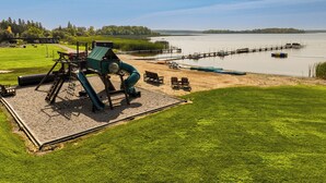 Un parque infantil con tobogán y estructuras para escalar, una zona de arena con bancos, y un lago con un muelle al fondo.