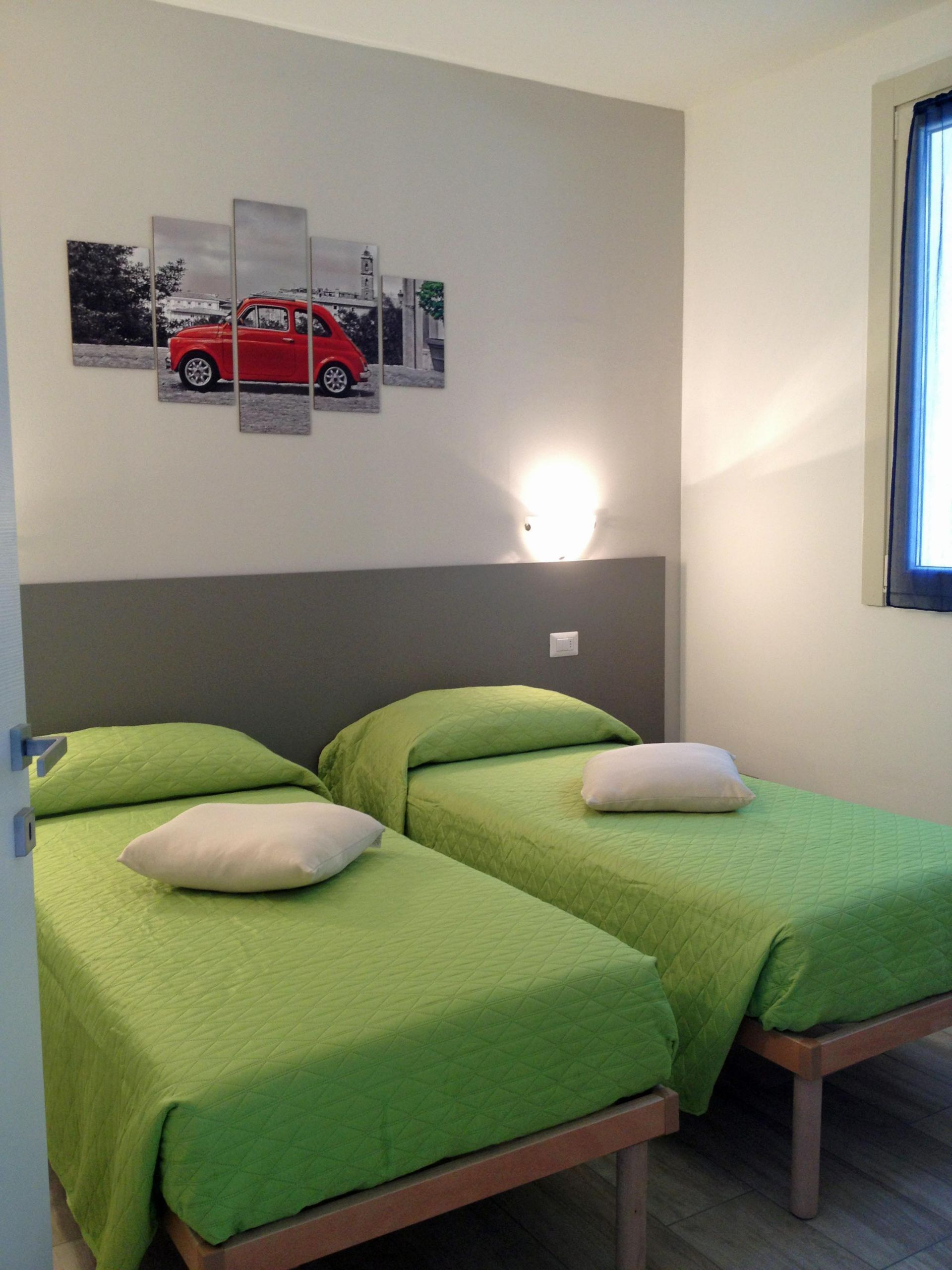 A room with two single beds, each with green bedding and white pillows, a grey headboard, and a wall-mounted light.