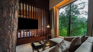 A modern living room with a large flat-screen TV, a sofa with cushions, a small table with a lamp, and a view of a forest through a large window. - Kasauli Hills Resort (Kasauli)