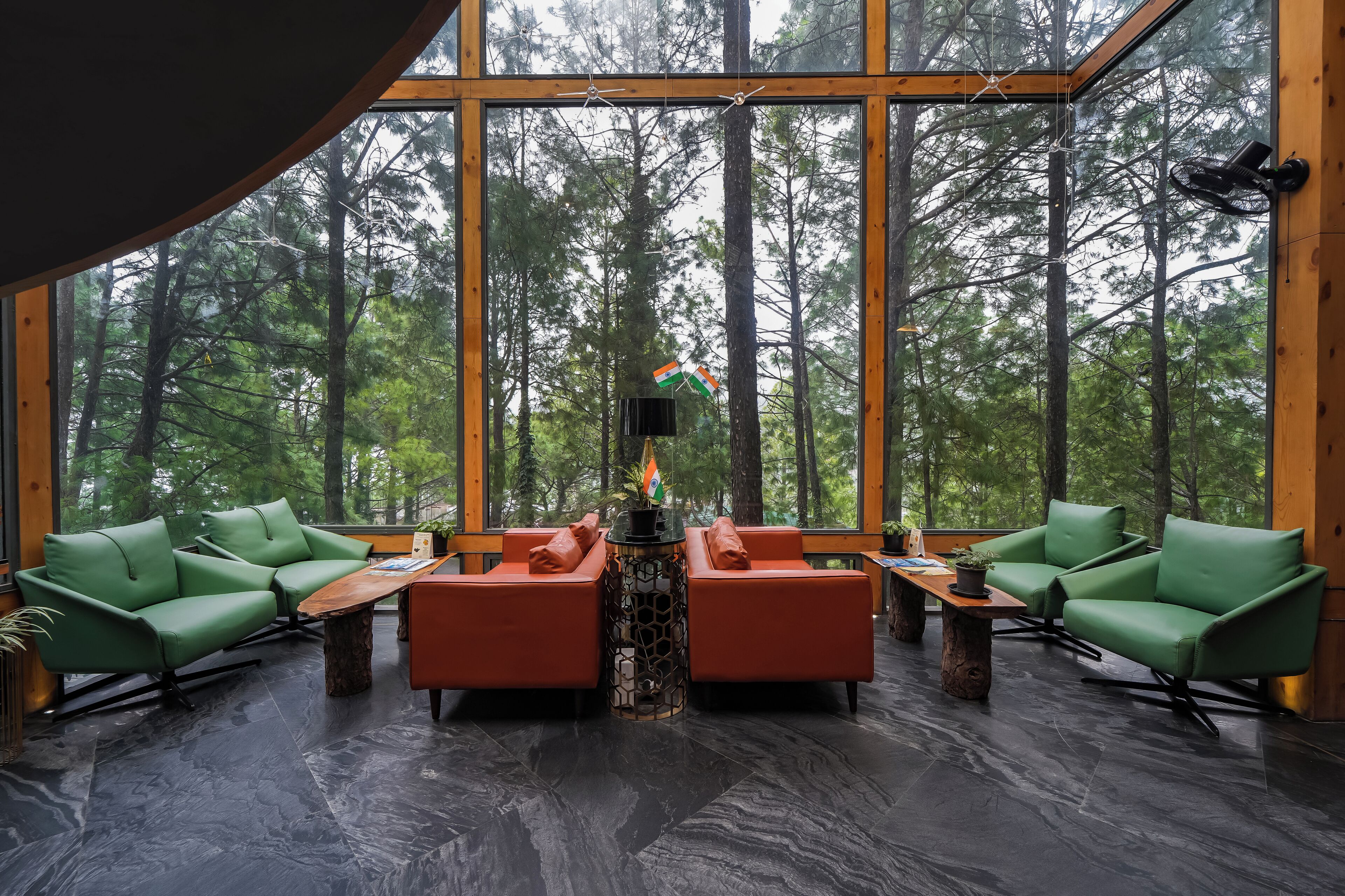Habitación amplia con ventanas grandes que dan a un bosque, una mesa central con sillas y una combinación de asientos verdes y anaranjados.