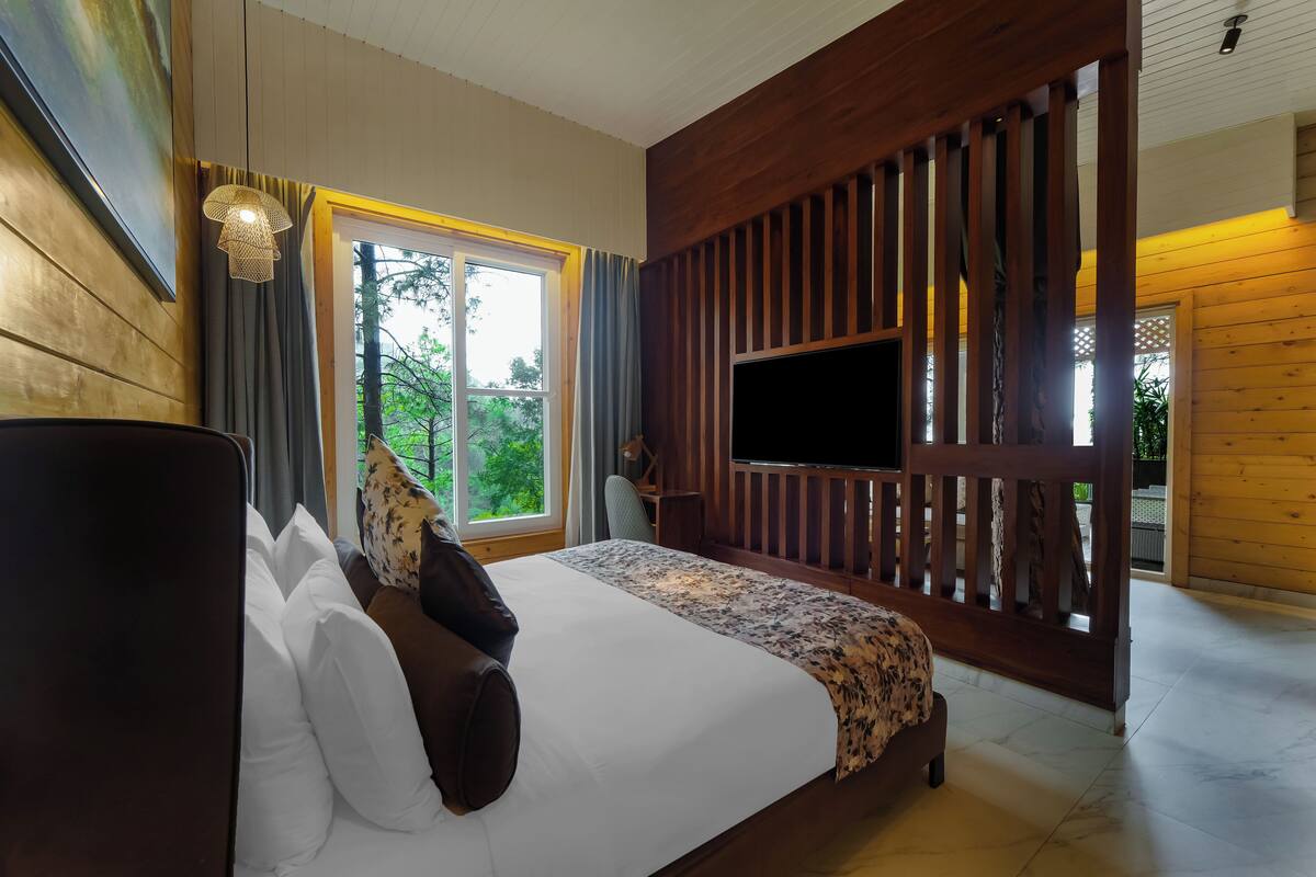 Quarto com uma cama grande, televisão fixada num painel de madeira, e vista para o verde lá fora.