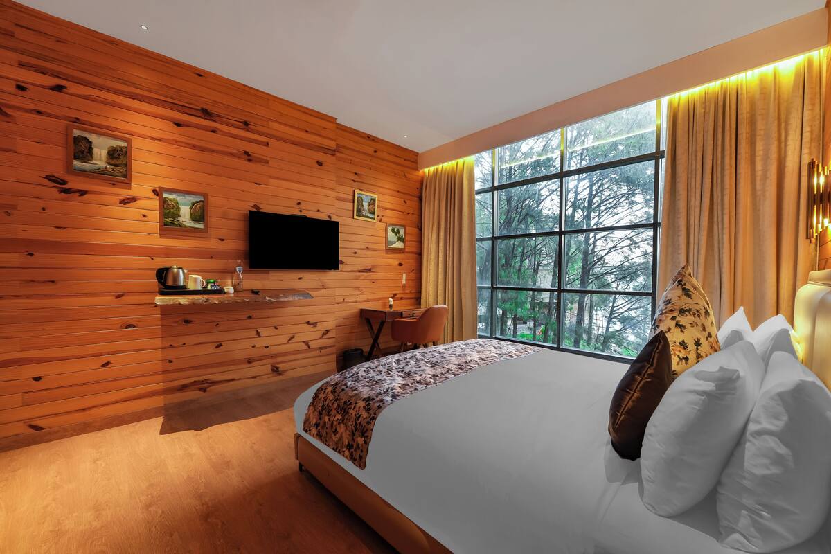 Quarto moderno com parede de madeira, cama com roupa de cama branca e estampada, escrivaninha com cadeira e uma janela grande com cortinas.