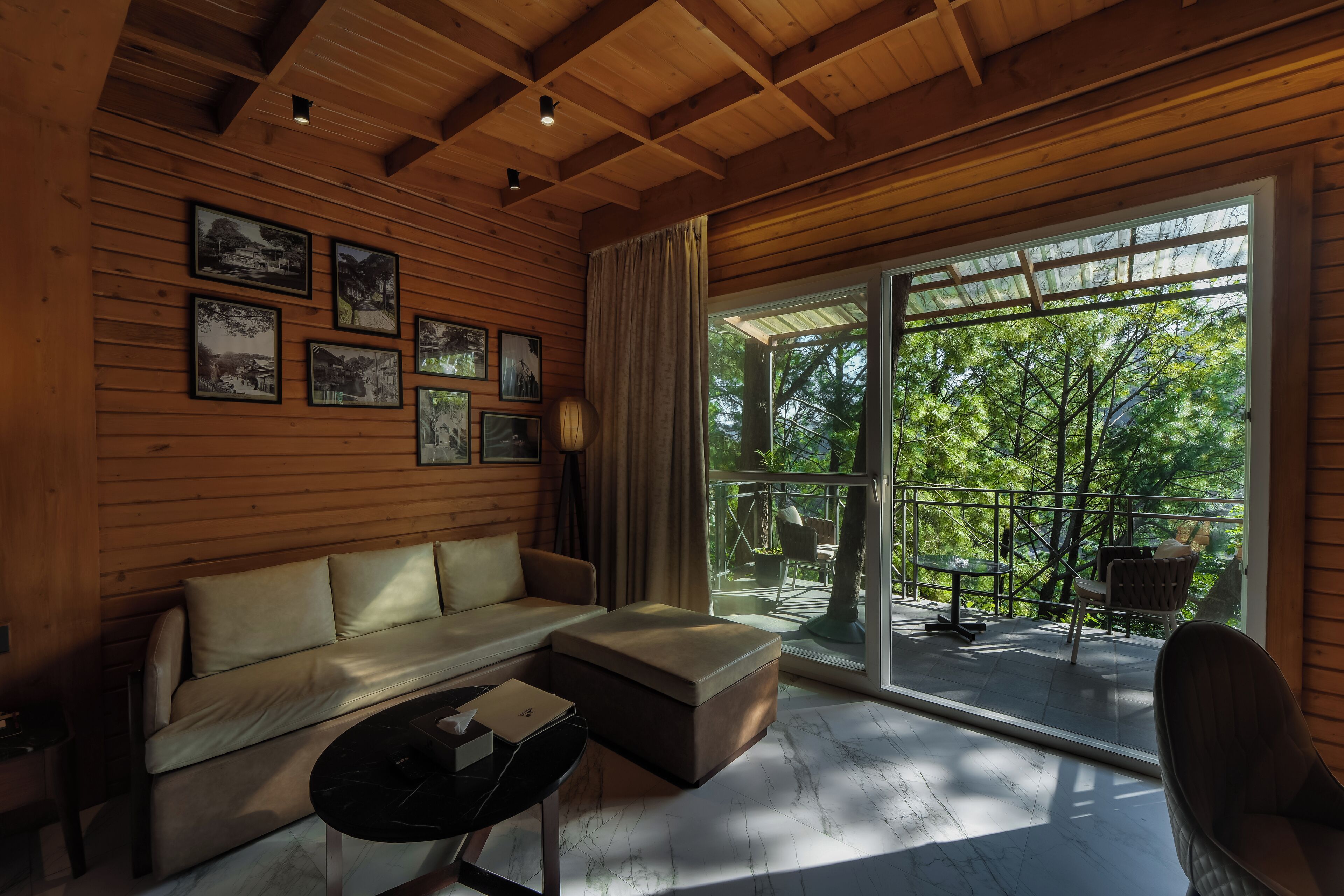 A cozy room with a wooden ceiling, a sofa, a coffee table, and a large sliding glass door leading to an outdoor area with a view of trees.