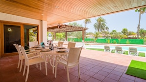 An outdoor patio with a wooden ceiling, white wicker furniture, and a view of a swimming pool and greenery.