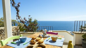 Une table en bois donnant sur la mer, une cafetière et diverses pâtisseries disposées sur un tapis bleu.