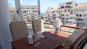 Un balcon avec une table en bois, deux chaises et une vue sur des bâtiments blancs et la mer.