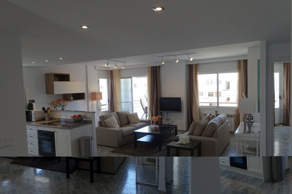 Una sala de estar moderna con sofá, mesa de centro y televisor, comunicada con una zona de cocina que incluye lavavajillas y armarios.