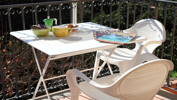 Une table en métal blanc sur laquelle se trouvent un livre et une tasse, un r écipient vert et un bol de collations, le tout sur un balcon.