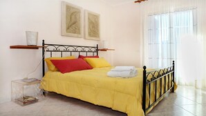 Un lit recouvert d’une couverture jaune, avec des oreillers rouges et jaunes, et une serviette blanche posée sur le côté. Deux tableaux encadrés sont accrochés au mur au-dessus du lit.