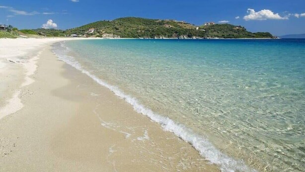 Ein Strand mit klarem türkisblauem Wasser, einem sandigen Ufer und einer Erhebung im Hintergrund mit Gebäuden.