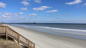 Une plage avec un escalier en bois descendant jusqu'au sable, une jetée visible au loin et un ciel dégagé.