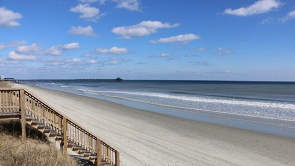 A beach with a wooden staircase leading down to the sand, a pier in the distance, and a clear sky.