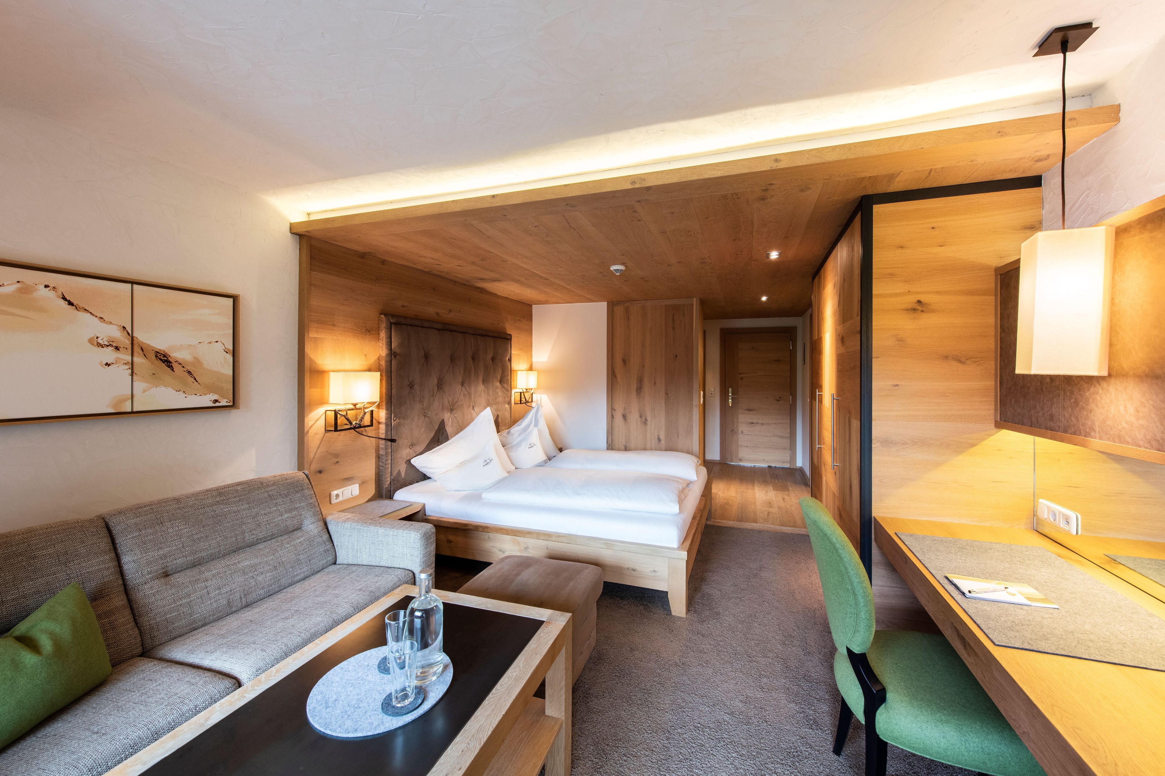 A hotel room with a bed, a sofa, a small table with glasses, and a desk with a chair.