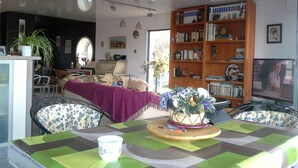 Un coin repas avec une table dressée pour deux personnes, entourée de chaises à motifs floraux. On aperçoit, au fond, une bibliothèque remplie de livres et d’objets décoratifs.