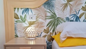 Ein ordentlich bezogenes Bett mit einem gelben und einem weißen Kopfkissen, ein Nachttisch aus Holz mit einer Lampe und eine Wand mit tropischem Blattmuster.