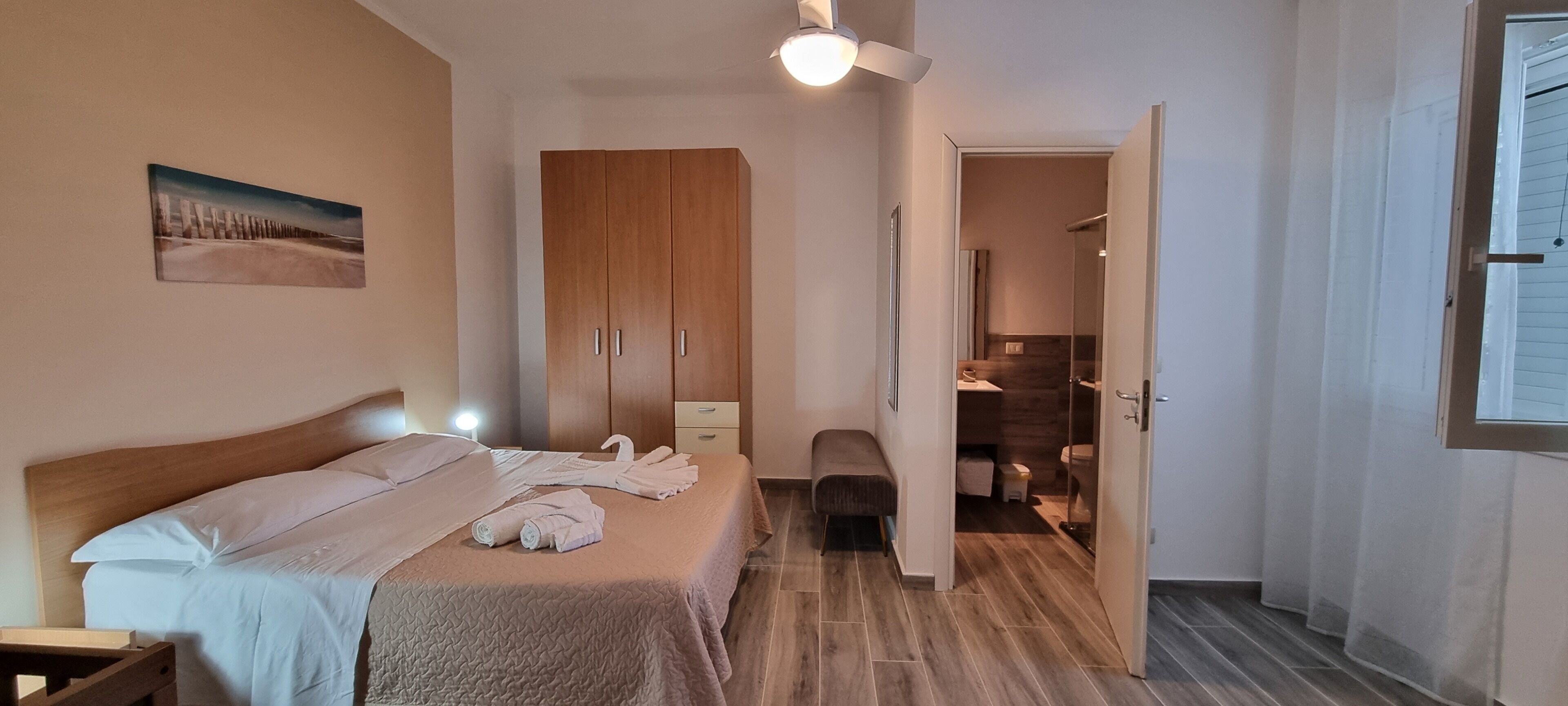 Ein Schlafzimmer mit Bett, Schrank, Bank und einem durchgehenden Türrahmen einsehbaren Badezimmer.