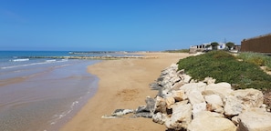 A sandy beach with a rocky shoreline, a clear blue sky, and a building with a blue roof in the background. - Comfortable villa with pool, close to the beach (scicli)