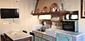 Una cocina con televisión montada en la pared, estufa a gas, horno y microondas. Hay una mesa de comedor con sillas y un estante sobre la estufa con platos y un reloj.