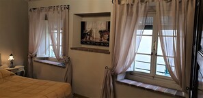 Un dormitorio con dos ventanas, una mesita de noche con lámpara y una cama con cubrecama acolchado.